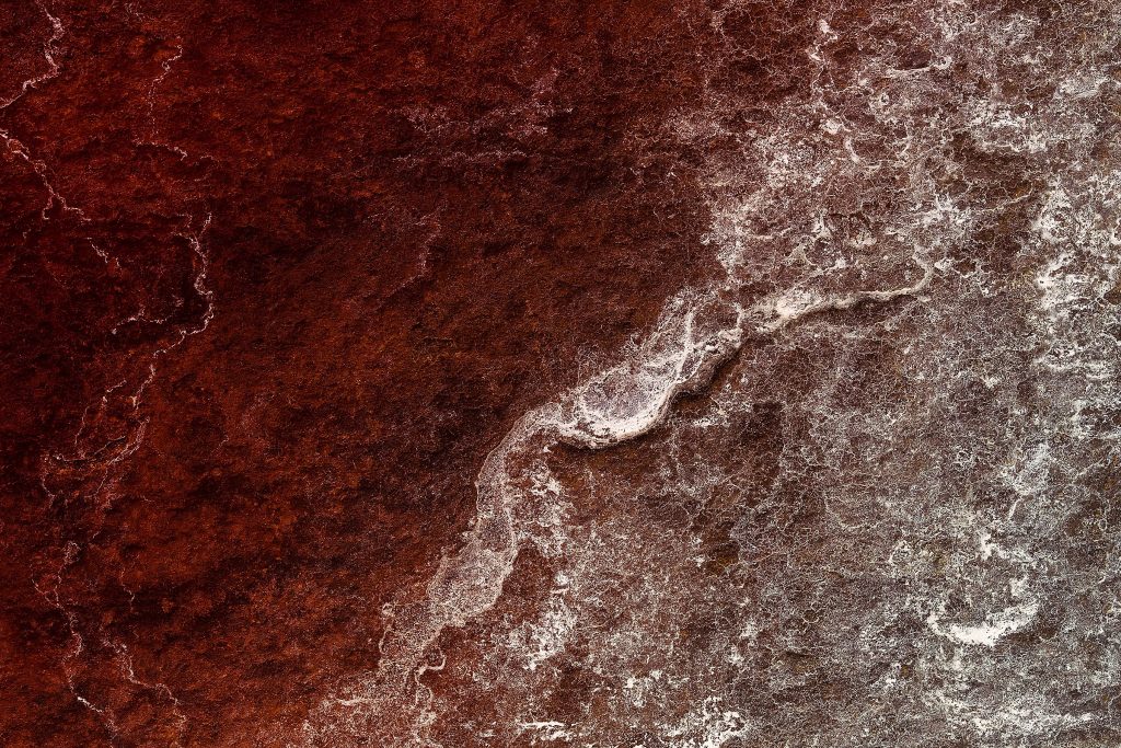 Close-up photograph of a basalt stone with white residue, creatively recolored in dark red to depict a sea of blood and rolling waves. Stoic stone forms evoke waves of blood, their silent presence echoed by dynamic lime deposits, whose froth and foam are rigid in time.