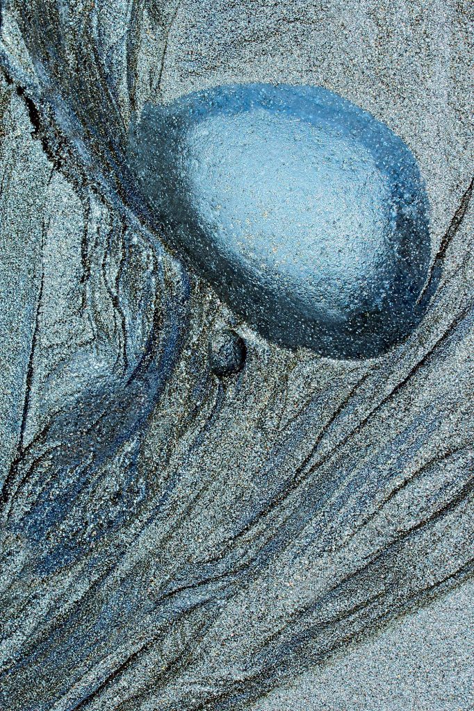 "DEFORMATION" - Close-up photograph of a round stone and sand on the beach of Bali, Indonesia, in a delicate silver-blue tone. The dynamics of ground movement. An abstract distortion of reality to which the shifts in deformation are linked.