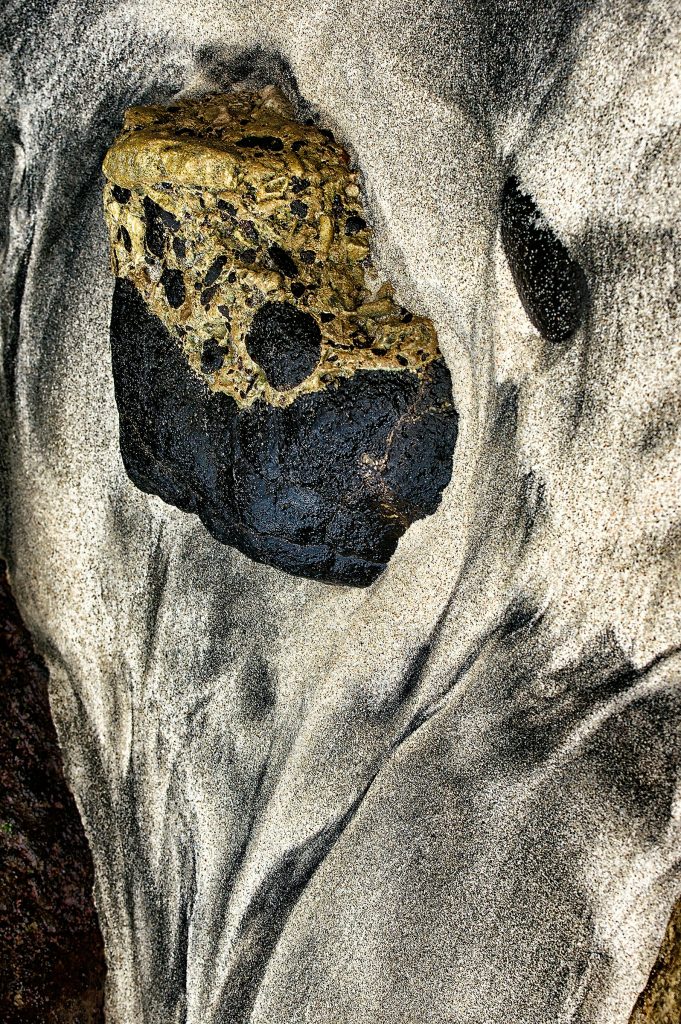 "Keeper of Gunung Agung" - Close-up photograph of a black and brown-white stone resembling a skull on a black and white beach, inspired by Balinese culture and Gunung Agung volcano.
