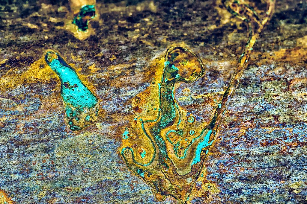"Longing": Abstract metal surface photograph featuring oxidized parts and shimmering hues of turquoise green and blue from copper patina. Two abstract shapes evoke a sense of longing and connection across the metallic landscape.