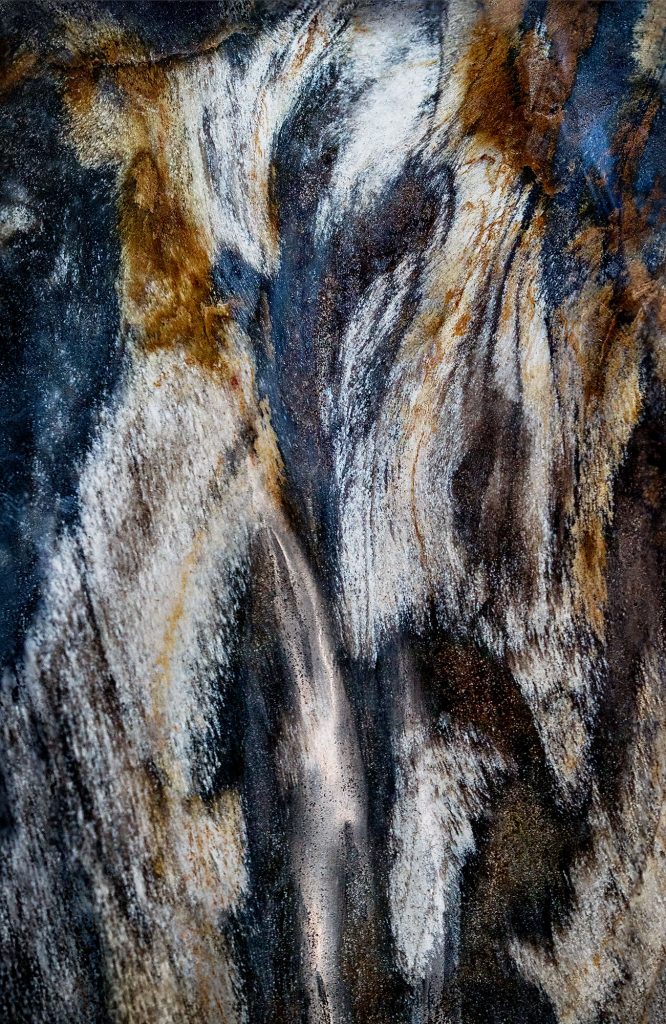"Spiritus": Close-up photograph of a polished petrified wood, featuring bluish and white tones with accents of wood brown, and depicting a small figure ascending, evoking themes of transcendence and serenity.